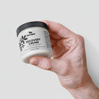 Hand holding a jar of recovery cream with a visible brand logo on a light background