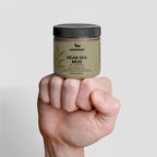 Hand holding a jar of Nestlève Dead Sea Mud on a gray background